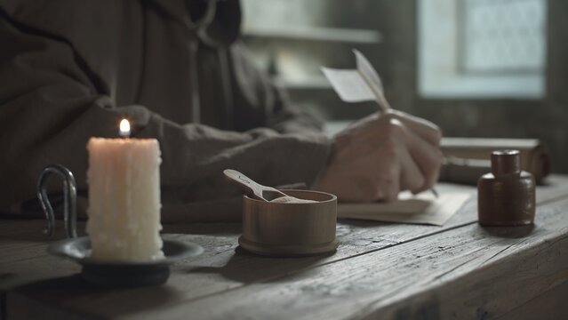 Pounce pot with a small spoon on a wooden table near a burning candle and a scribe writing a manuscript in background inside a scriptorium
