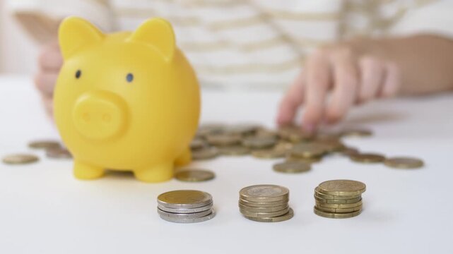 Yellow piggy bank stands beside several coin stacks while hand rests near table edge. Future savings deposit concept shows organized wealth building through consistent contribution