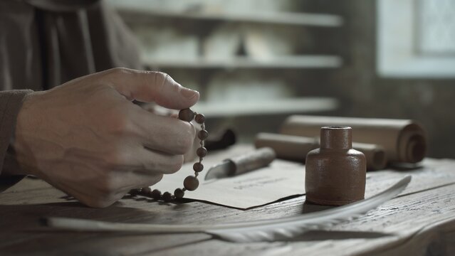 Hands of a scribe praying with rosary beads next to a ceramic inkwell and a white quill pen on a wooden table inside a medieval scriptorium