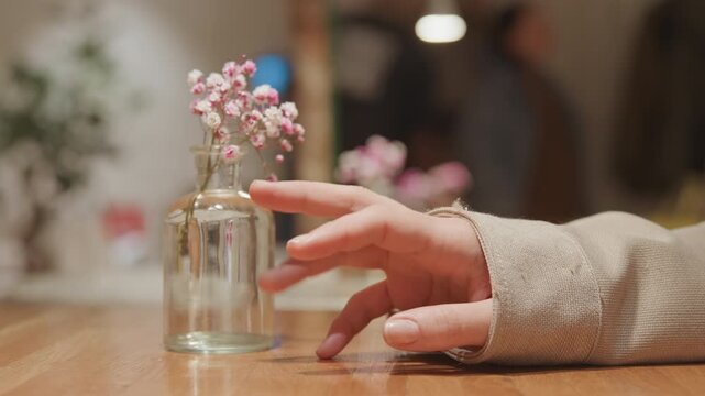Close up of hand resting on wooden table near clear glass vase with pink flowers. Background shows blurred interior space with soft lighting and additional floral arrangements. Hand remains