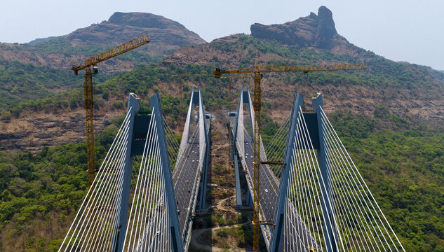Aerial view of the Missing Link Suspension Bridge under construction with tall cranes, cable-stayed structures, and highway traffic amidst lush green hills in Lonavala, Maharashtra, India.
