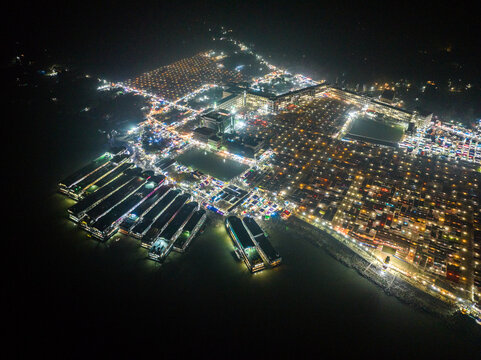 Aerial view of the illuminated Charmonai Mahfil gathering at night, featuring numerous tents, docked boats along the riverbank, and vibrant lights in Charmonai, Barisal Division, Bangladesh.