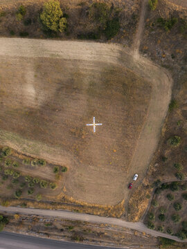 Aerial view of a cross-shaped marker in a dry field with parked cars and olive groves in the rural landscape of Gravina in Puglia, Apulia, Italy.