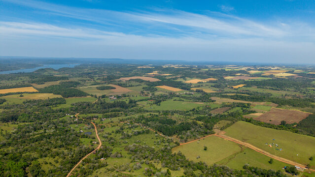 Aerial view of rolling rural landscape with patchwork fields, dirt roads, forest patches, and a distant river under a clear blue sky in Nueva Alborada, Itapua Department, Paraguay.
