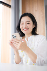 自宅でスマホを見ながら笑顔を見せる50代女性、家事の合間にネットやSNSを楽しむライフスタイルイメージクローズアップ考える © kapinon