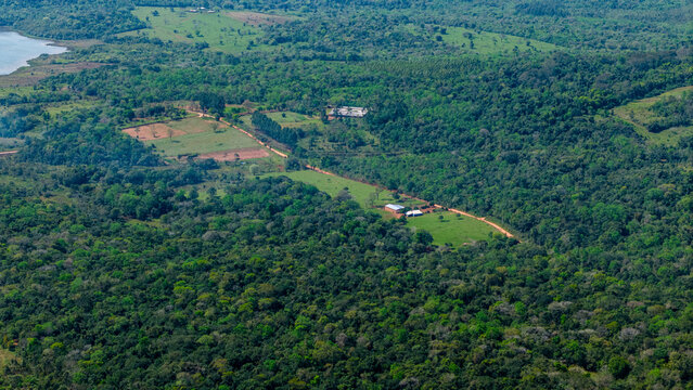Aerial view of a rural landscape featuring dense green forests, open grassy fields, and a dirt road connecting small farm buildings in Nueva Alborada, Itapua Department, Paraguay.