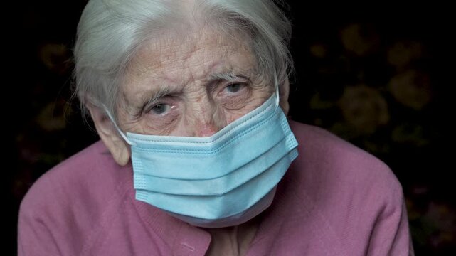 Sad elderly woman with grey hair wearing protective surgical face mask and looking directly at camera. Old granny female wrinkled face expressing sorrow and loneliness during the pandemic