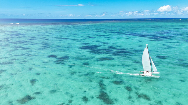 Aerial view of a small sailboat gliding through crystal clear turquoise lagoon waters with coral reefs visible beneath the surface in Trou-aux-Biches, Pamplemousses District, Mauritius.