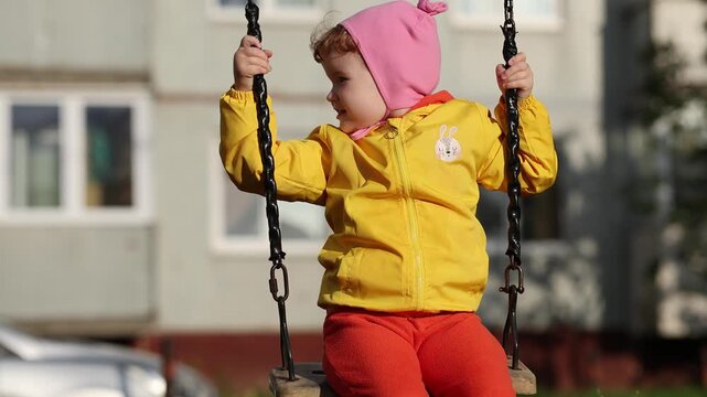 Little girl in pink hat and yellow jacket swinging on swing at playground. Happy child playing outdoors in autumn. Childhood, joy and family leisure activity concept