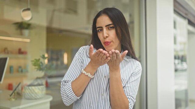 Young woman in a striped shirt blowing a kiss with cupped hands at a shop storefront window on a street, smiling through glass reflection; playful affection.