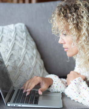 Woman with curly hair is concentrating on her laptop, typing and browsing, performing remote work or studying from the comfort of her home sofa with a casual and relaxed posture