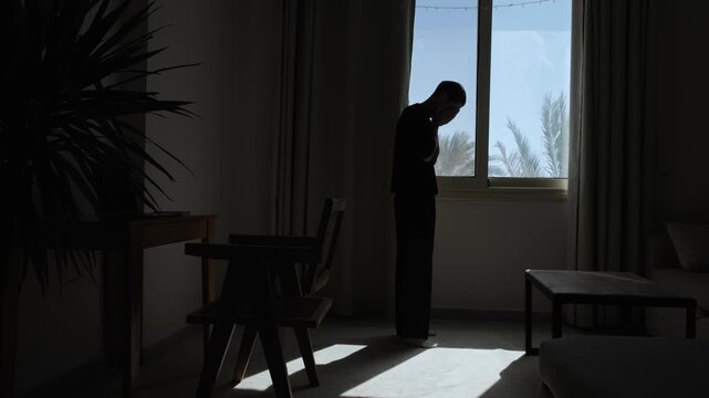 Silhouette of a distressed man standing alone by a window and covering his face, overwhelmed by emotional stress, worry and a difficult realization in a dark dramatic room