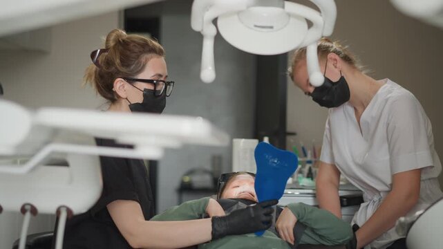 Close up of two dental professionals attending to young patient during orthodontic procedure. One assistant holds blue oral retractor while another observes with medical equipment visible