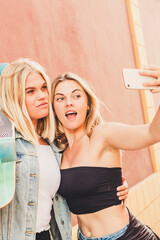 Two smiling blonde friends take a selfie with a smartphone, sharing summer joy and a skateboard in...