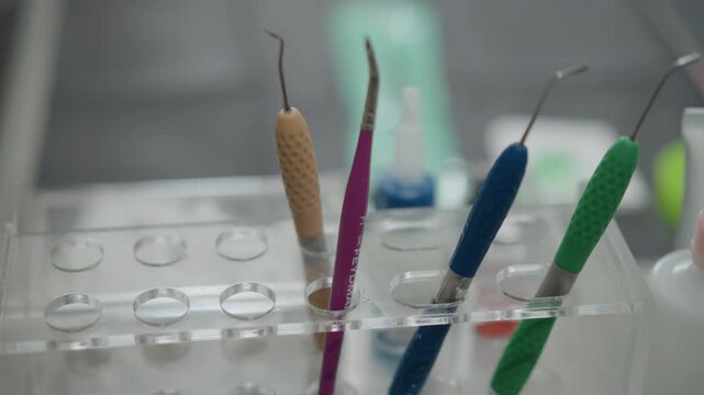 Close up of dental instruments including scalers and probes in clear plastic holder. Instruments are color coded with beige pink blue and green handles visible against out of focus clinical