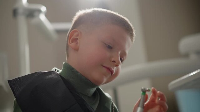 Close up of young boy examining dental tool while wearing protective bib in clinical setting. Boy holds small instrument observing it closely as soft light illuminates scene with medical
