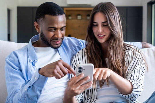 Young diverse couple relaxing at home on a couch, using a mobile phone together and interacting with content, representing connection, digital lifestyle, and shared leisure time