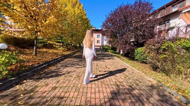 Young blonde woman walking along a scenic path with vibrant autumn trees and clear blue sky, back view of slender girl enjoying sunny fall day, outdoor lifestyle concept