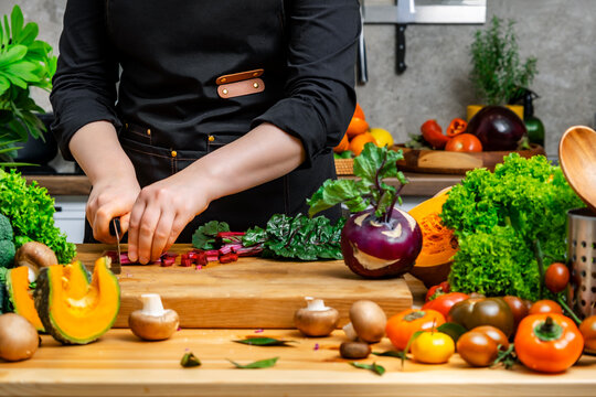 Cook prepares healthy vegetarian meal chopping fresh swiss chard leaves on wooden board. Chef in black apron cuts raw organic ingredients for vegan dinner in modern kitchen.