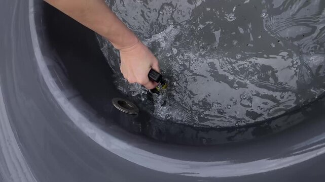 Person uses plunger cleaning clogged jacuzzi bathtub drain. Water flows inside dark hot tub during plumbing maintenance service. Professional worker unclogs pipe ensuring clean home sanitation.