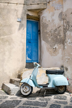 Procida, Italy - August 7, 2026. Vintage Blue Scooter Parked in Front of a Weathered Blue Door in Procida. A charming, rustic view of a classic light-blue vintage scooter parked on a dark cobblestone 
