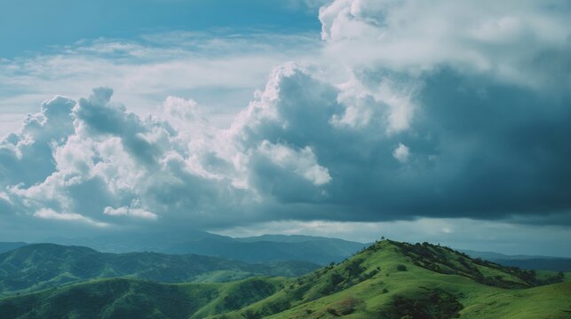 Lush green rolling hills and mountains under a dramatic, cloudy blue sky. A scenic natural landscape showcasing outdoor beauty and vastness.
