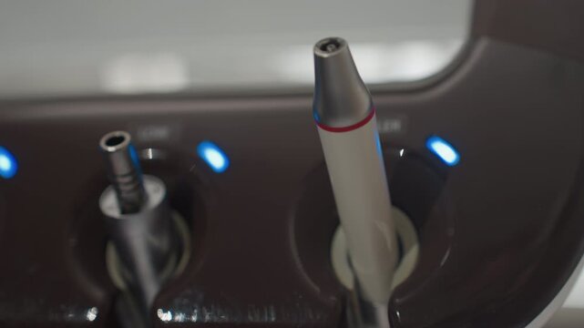 Close up of dental handpieces in docking station with blue indicator lights. instruments are stationary showcasing their metallic and plastic components against dark brown base. Soft