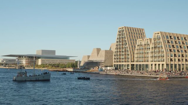 Boats cruise past modern luxury apartments on Paper Island in Copenhagen harbor. Tourists relax on the sunny waterfront near the Opera House. Scandinavian architecture highlights elite living.