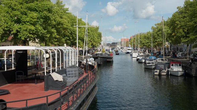 Houseboats and motorboats moor along a scenic canal in Copenhagen. Green trees shade the historic waterfront promenade. Danish nautical culture highlights peaceful European city vacation.