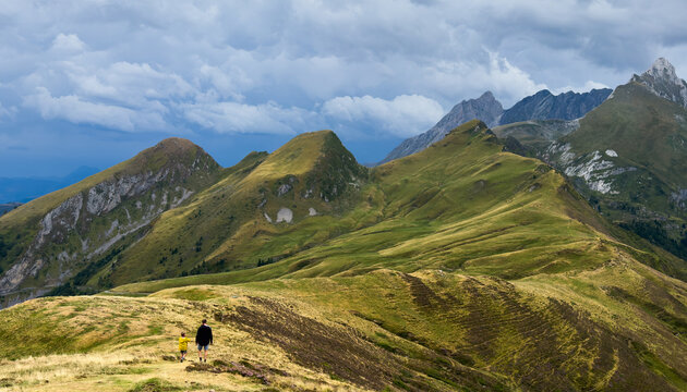 two people hiking on a mountain trail in a peaceful, adventurous landscape with green grassy slopes and rocky peaks under a cloudy sky from a higher vantage point.