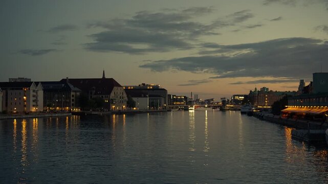 Illuminated buildings reflect on a calm harbor canal in Copenhagen at twilight. City lights glow along the urban waterfront during dusk. Evening scenic view conveys peaceful travel atmosphere.