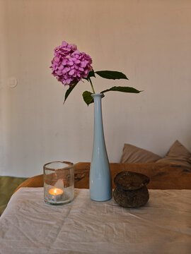 Minimalist composition featuring a pink hydrangea in a slender tall blue vase alongside a small burning candle and stacked smooth balance rocks on a wooden table with linen cloth.