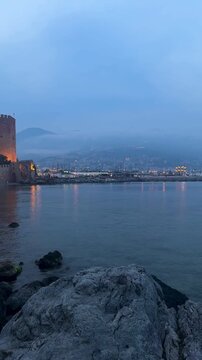 Alanya, Turkey, Kizil Kule Red Tower at dusk with calm sea, rocky shore, and glowing city lights under misty mountains
