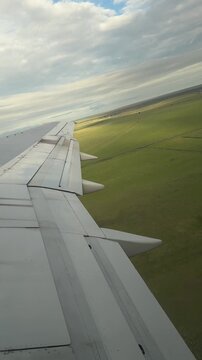 View from airplane window of wing taking off over green field in overcast summer weather. Clouds float in the sky while the shadow of the flying airplane is visible on the ground