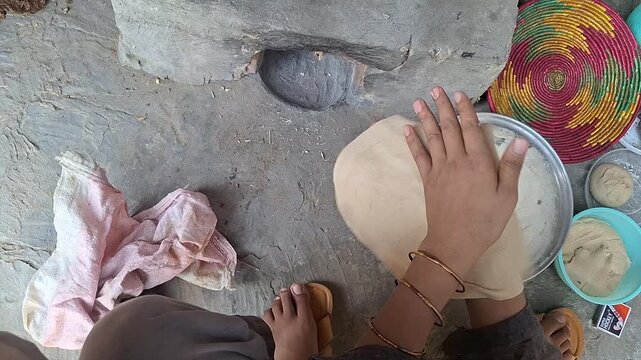 Rural woman hand tossing and flattening wheat flour dough to bake traditional roti flatbread beside an earthen stove in a village kitchen   