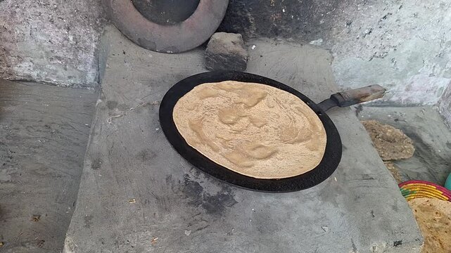 Applying clarified butter ghee with a spoon onto traditional paratha flatbread baking on an iron tawa pan over a mud stove   