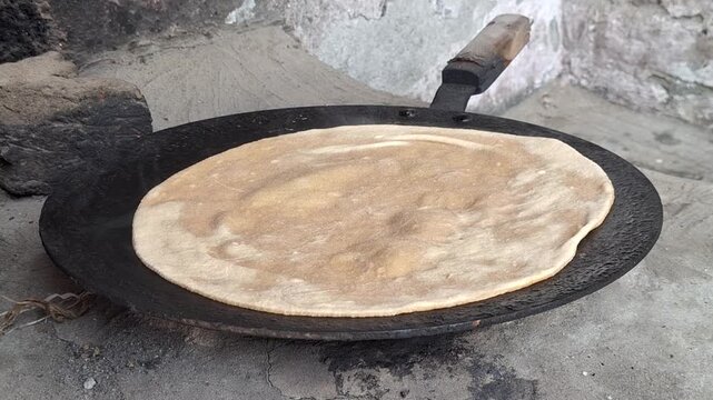 Traditional flatbread paratha or roti cooking on a rustic iron tawa griddle over an open fire flame with subtle sparks and warm glowing coals in an authentic rural kitchen setting