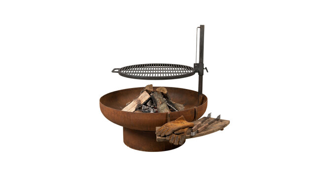 Rustic metal fire pit with burning wood logs and a suspended grill grate sits next to leather gloves and cooking tools on a grey background.