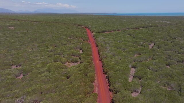 A cinematic aerial forward-tracking shot following a striking red-soil road cutting through dense New Caledonian bushland, with the distant coastline faintly visible on the horizon. This scene has str