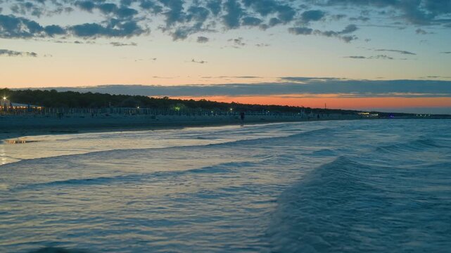 Beautiful beach at sunset with ocean waves rolling onto the shore. Serene coastal landscape during twilight with a vibrant orange horizon, dramatic clouds, and the silhouette of a beach resort in the