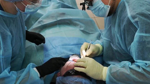 Dental surgeon and female dental assistant perform dental implant procedure, with assistant using saliva ejector while dentist works on patient in chair wearing sterile gown, gloves and cap.