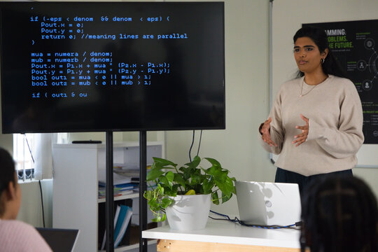 Indian woman wearing sweater teaching African American teens coding on TV, laptop, plant