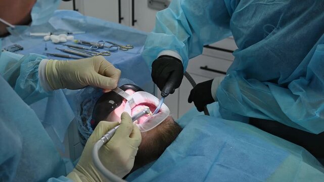 Dental surgeon and female dental assistant perform dental implant procedure, with assistant using saliva ejector while dentist works on patient in chair wearing sterile gown, gloves and cap.