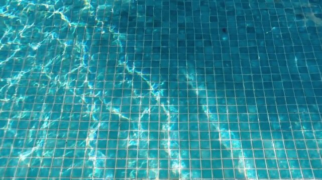 Slow motion close up of pool water rippling over mosaic tile floor with sunlight reflections in outdoor swimming pool during bright summer daytime calm mood
