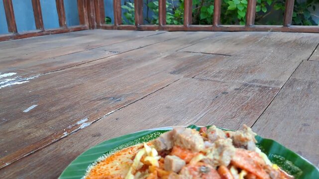 Traditional Tajhin Sorah Madura porridge served on banana leaf plate at joglo wooden table. Authentic Indonesian halal cuisine with coconut milk, tofu tempe sambal, egg, meat, peanuts. 4K food stock.