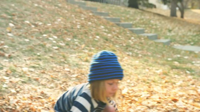 Boy in a striped beanie walking along a path and sitting down in a pile of fallen leaves