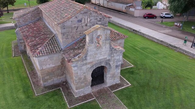 Elevated high-angle drone orbital arc over the tiled roof layout of San Juan de Ba&ntilde;os Visigothic church, asymmetrical framing on cloudy day.