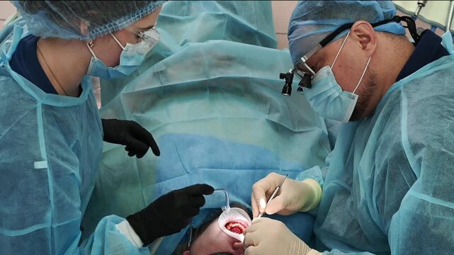 Dental surgeons performing gum restoration and dental implant placement, wearing sterile gowns, masks and gloves, working inside patient mouth during oral surgery in clinic
