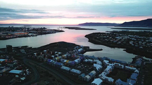 Aerial panorama of Reykjavik harbor and waterfront homes during sunset, with calm coastal water, pastel sky and distant Mount Esja on the horizon 4K.