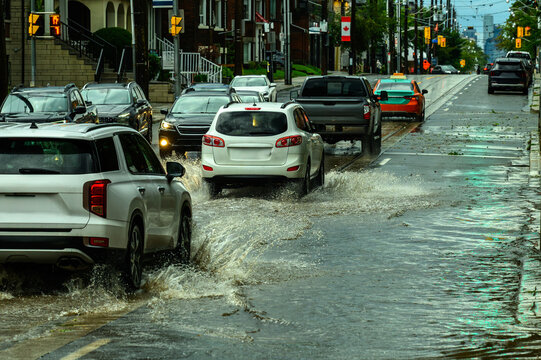 Traffic navigates a flooded city street lined with parked cars and residential buildings after a storm